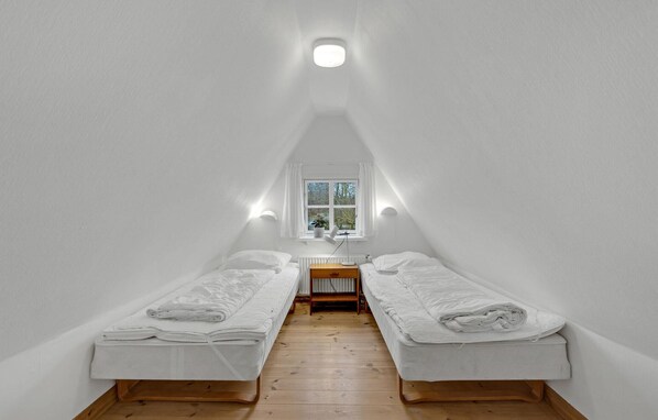 A small, white, attic-style bedroom with two single beds, a wooden floor, a window with a view, and a ceiling light.
