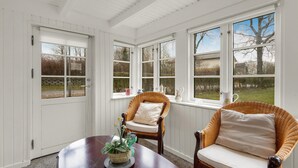Ein Zimmer mit weißen Wandverkleidungen, einem großen Fenster mit Blick auf Bäume und einen Garten, zwei Korbstühle mit Kissen und einem runden Holztisch mit einer Pflanze.