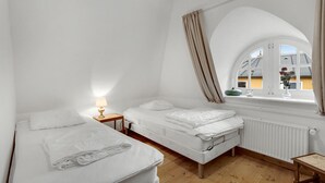 Ein kleiner, weißer Raum mit zwei Betten, einem Nachttisch mit Lampe, einem Fenster mit Vorhängen und einer Heizung.
