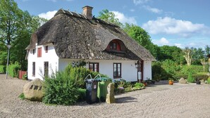 Ein traditionelles Haus mit Strohdach, weißer Fassade und roten Fensterrahmen, umgeben von viel Grün und einer Schotterauffahrt.