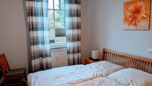 Une chambre à coucher comprenant un lit, une chaise, une table de chevet, un radiateur et une fenêtre avec des rideaux.