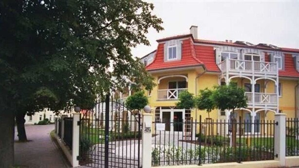 Un bâtiment jaune avec un toit rouge, une entrée fermée par un portail et un petit jardin.