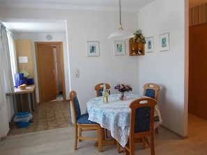 Et spiseområde med et bord dækket til fire personer, omgivet af træstole. Der er en vase med blomster på bordet, og en hængende lampe over.