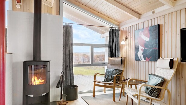 Ein gemütliches Zimmer mit Kamin, einem großen Fenster mit Vorhängen, einem Bild an der Wand und einer Sitzecke mit Tisch und Stühlen.