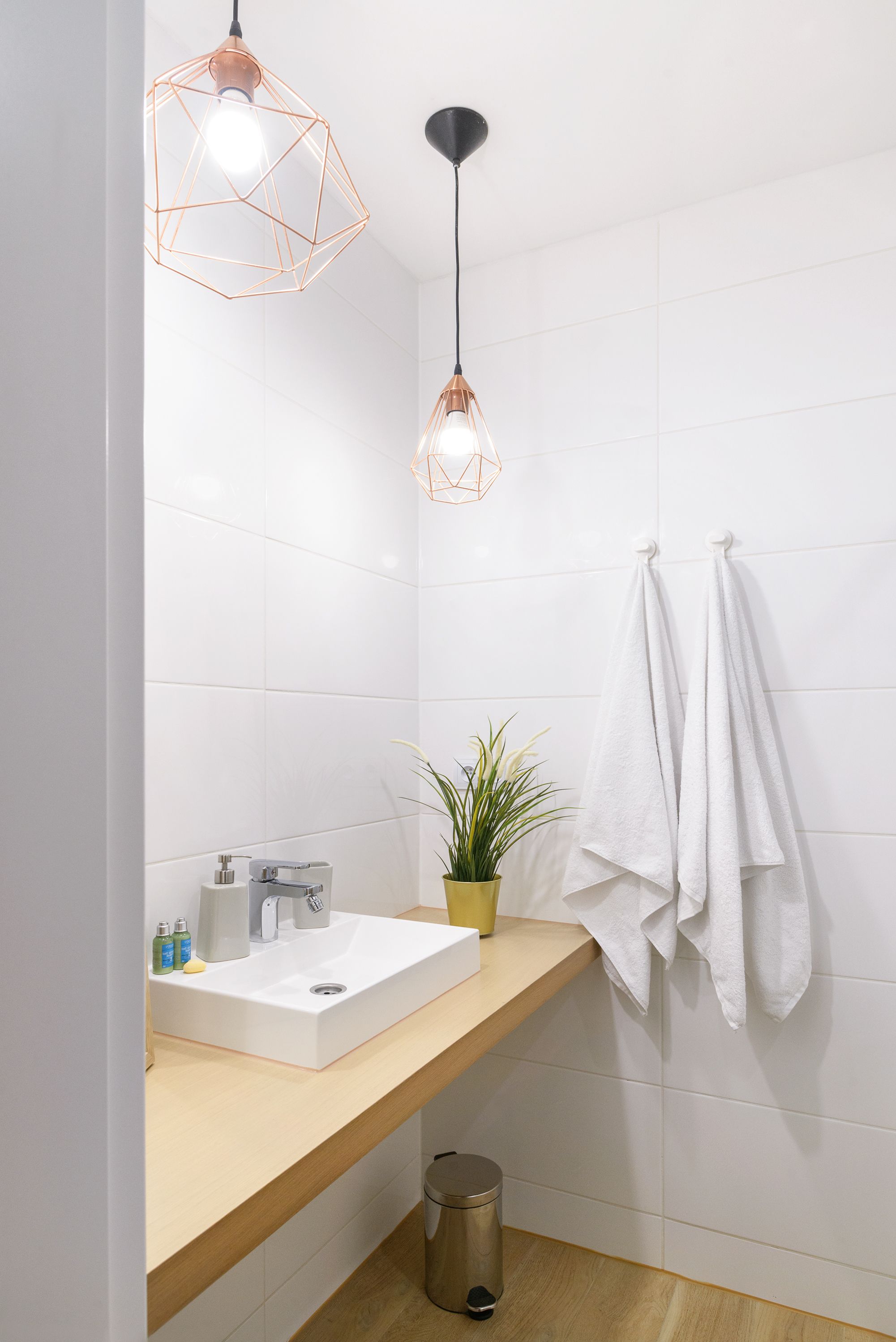 A modern bathroom with a white sink, two geometric pendant lights, a potted plant, and two towels hanging on a rack.