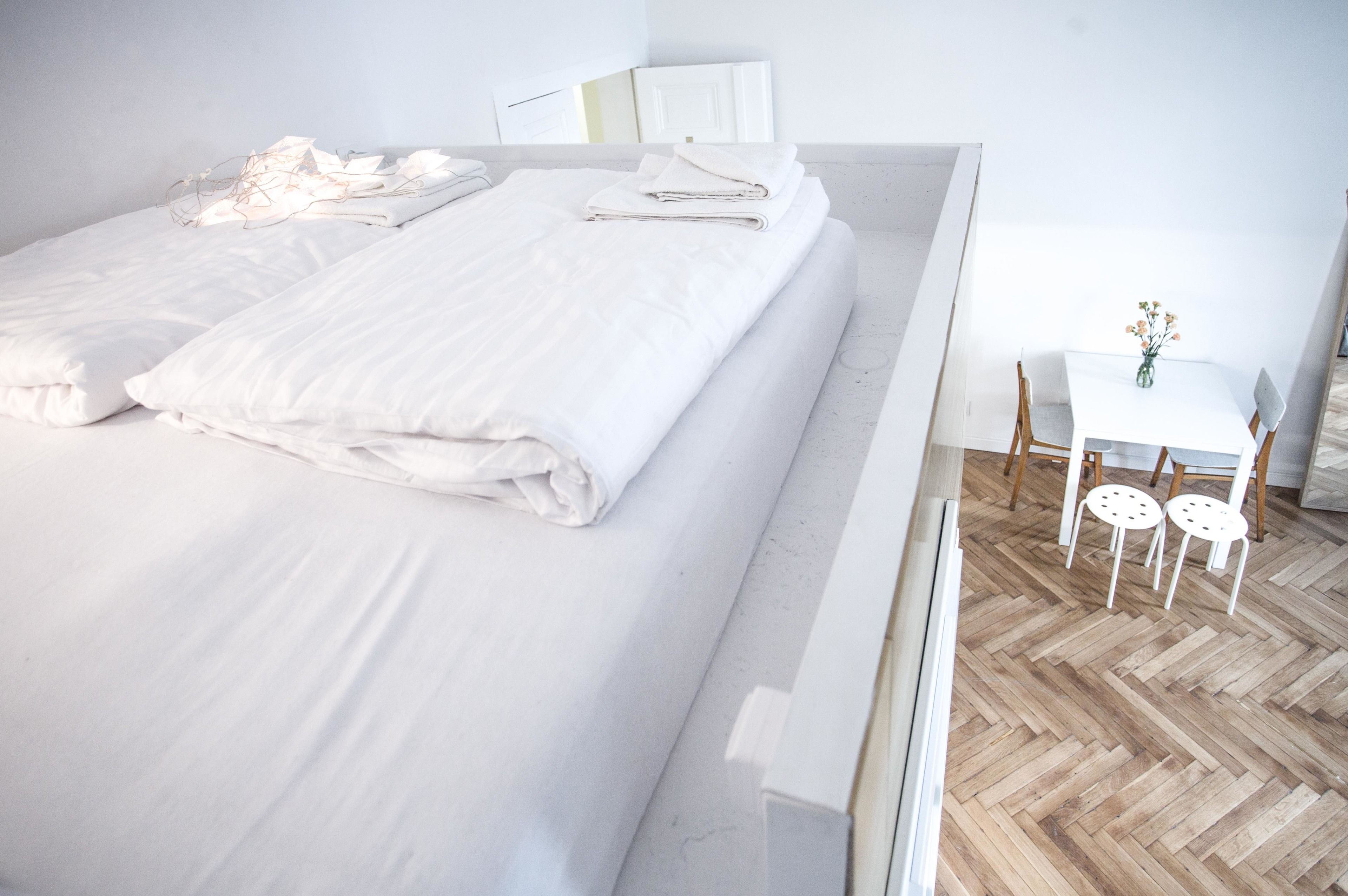 A neatly made bed with white linens, a small table with chairs, and a vase with flowers.