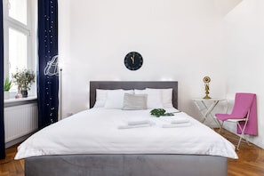 A neatly made bed with white linens and towels, a wall clock, a small table with a lamp, and a pink chair.