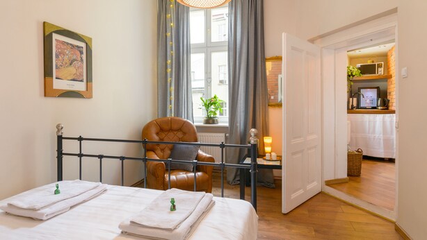 Un dormitorio con cama, una silla, una mesita de noche con lámpara, una ventana con cortinas y un cuadro enmarcado en la pared.