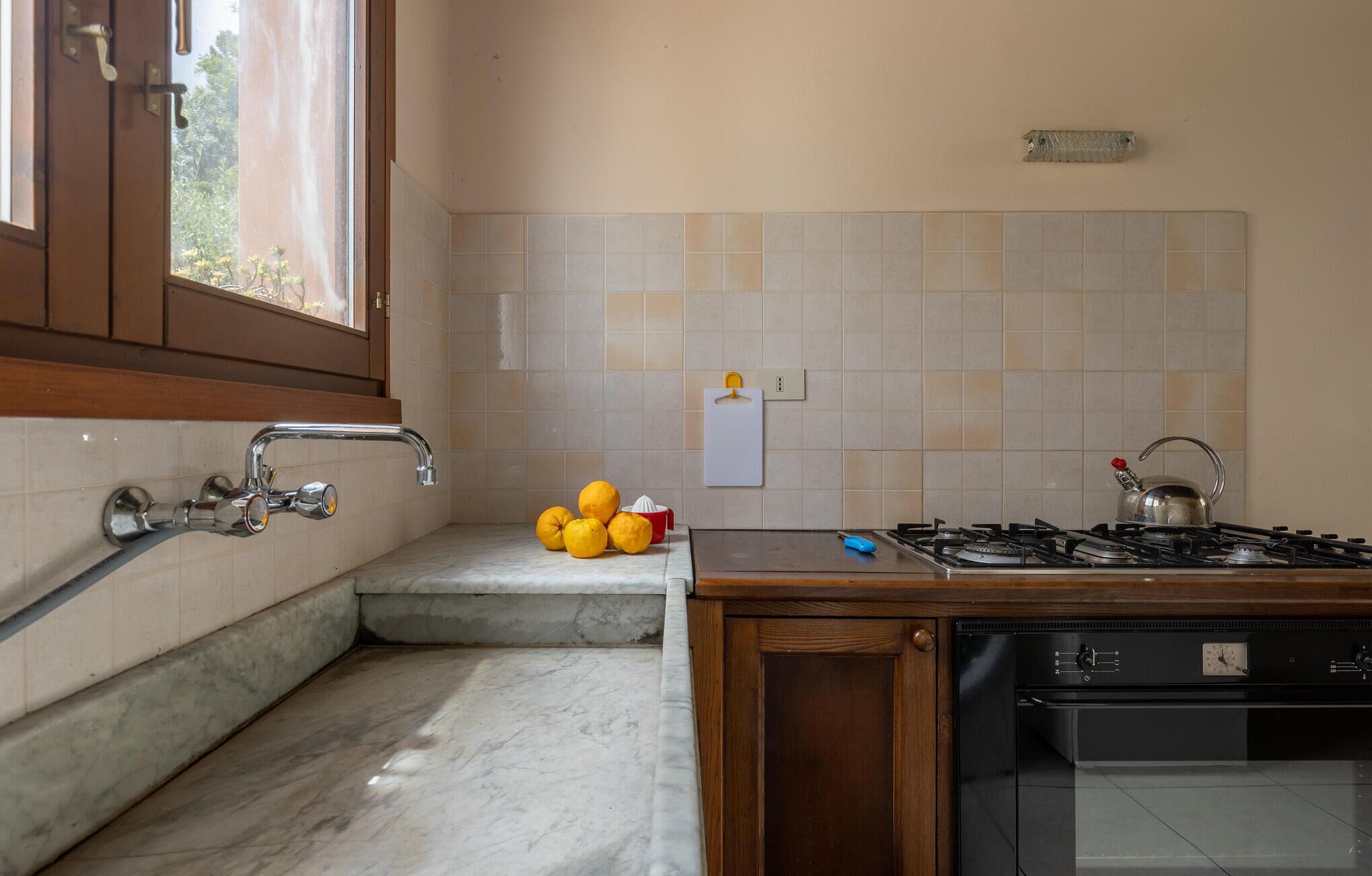 A kitchen with a gas stove, oven, and a sink with a faucet. There are lemons on the counter and a window with a view of trees.