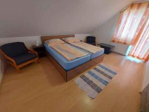 Een slaapkamer met een bed, een stoel, een nachtkastje, een bedtafel en een raam met gordijnen.