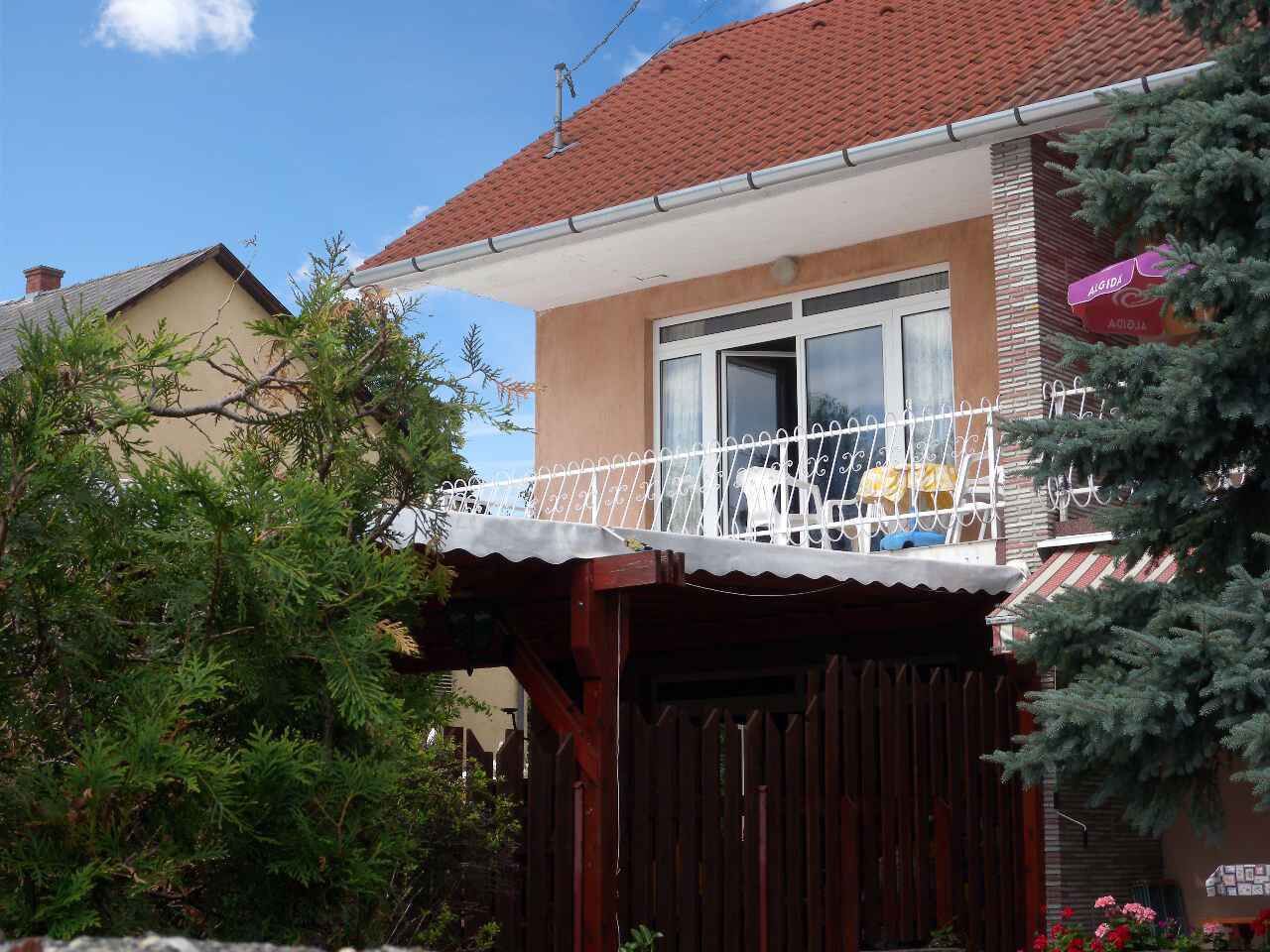 Ein zweistöckiges Haus mit rotem Ziegeldach, einem Balkon mit weißen Geländern und einem hölzernen Zaun davor.