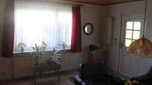 Een woonkamer met een zwarte leren bank, een houten tafel met planten, een radiator en een raam met rode gordijnen.