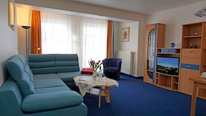Ein Wohnzimmer mit einem türkisblauen Sofa, einem kleinen Tisch mit einer Vase und einer Flasche, einem blauen Sessel, einem Fernseher und einer Holzkommode.
