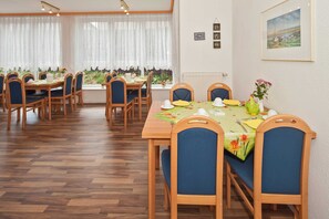 A dining area with wooden tables and chairs, a view of greenery through large windows, and framed pictures on the wall.