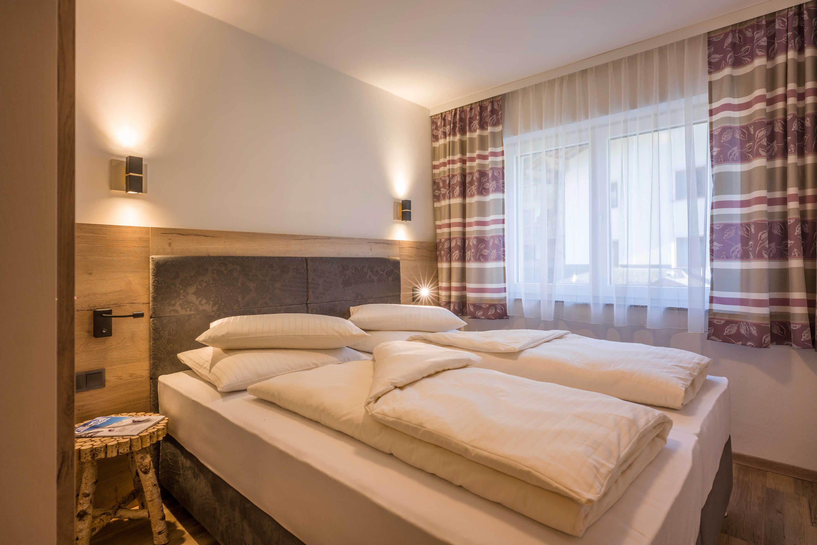 Quarto de hotel com cama, abajures, janela com cortinas e um banquinho.