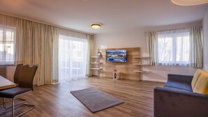 A modern living room with a flat-screen TV, wooden flooring, a sofa, a dining table with chairs, and a large window with curtains.