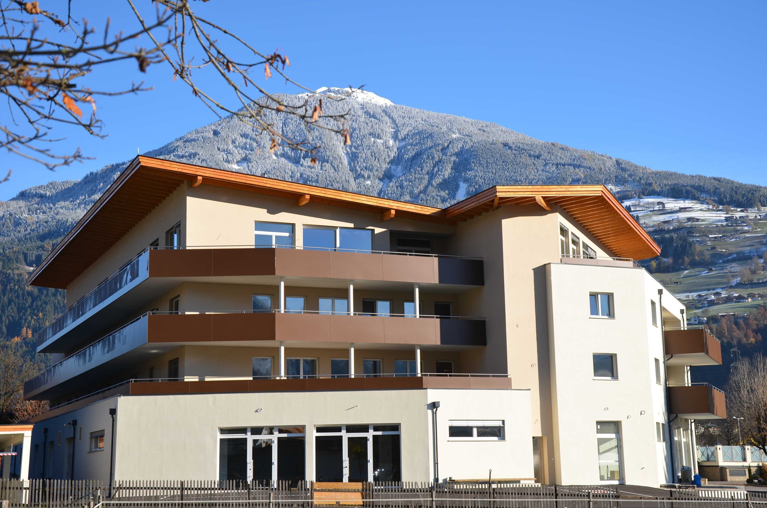 Un edificio de varios pisos con balcones, techo plano y montañas al fondo.