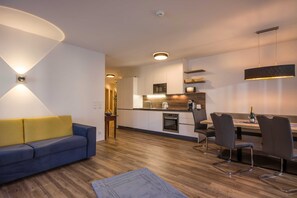A modern living room with a sofa, a dining area with chairs, a kitchen with appliances, and a ceiling light fixture. - Apartment Dorfplatzl - Aparthotel **** AlpTirol (Kaltenbach)