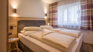 A hotel room with a bed, bedside lamps, a window with curtains, and a small stool.