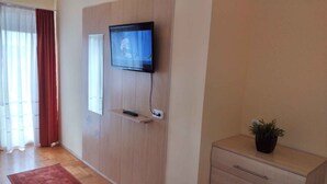 Een kamer met een schuifdeur, een wandtelevisie, een klein plankje met een afstandsbediening, een potplant en een houten dressoir.