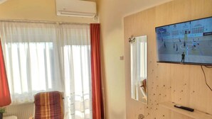 Een woonkamer met een aan de muur bevestigde televisie, een stoel, een bijzettafeltje en een raam met gordijnen.