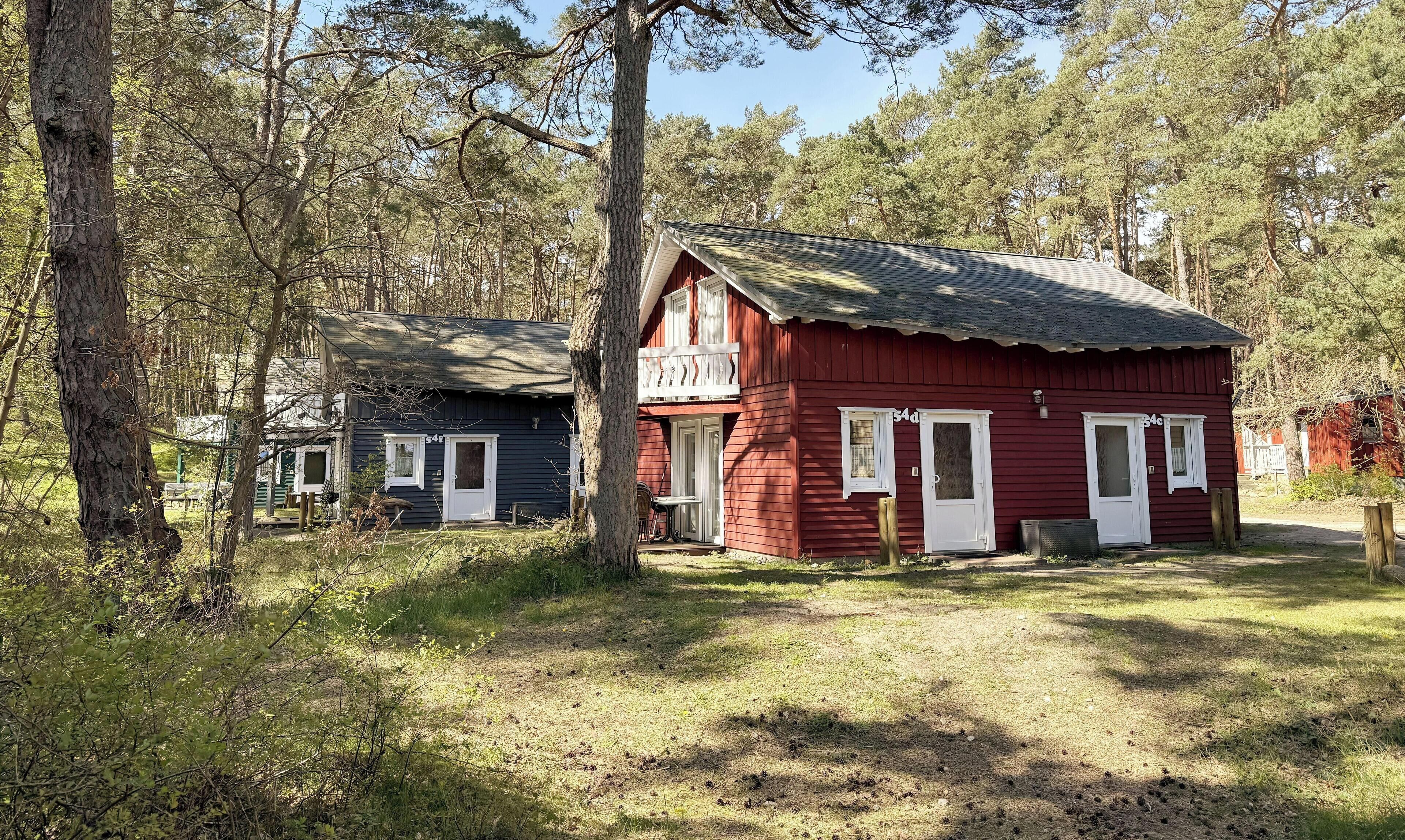 Two cabins in a forested area, one red and one dark blue, with a small porch and a door visible.