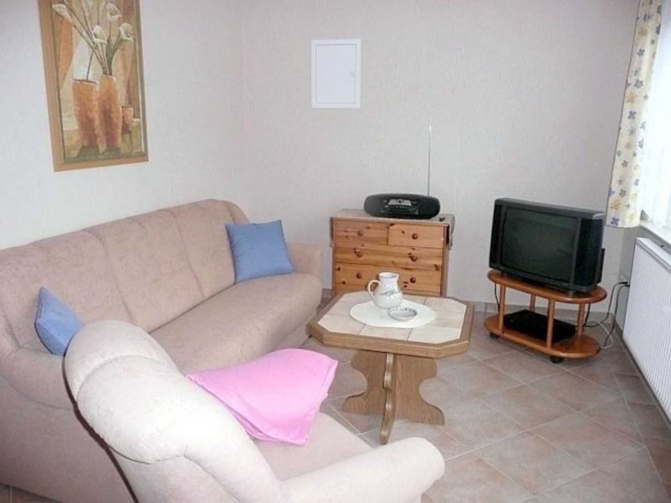 Ein Wohnzimmer mit einem beigen Sofa, einem kleinen Holztisch mit einer weißen Kanne, einem Fernseher auf einem Holzschrank und einer Heizung.