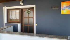 Entrada de un hotel con puerta de madera, ventana con cortinas de encaje, mostrador de granito y un cuadro en la pared.