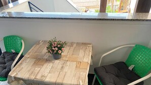 Una pequeña mesa al aire libre con una planta en maceta, dos sillas verdes y una barandilla blanca.
