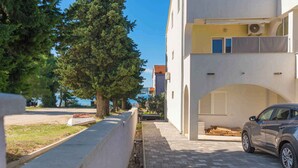 A building with a paved driveway, a parked car, and a large tree in the foreground. - Holiday apartment with air conditioning (Biograd na Moru)