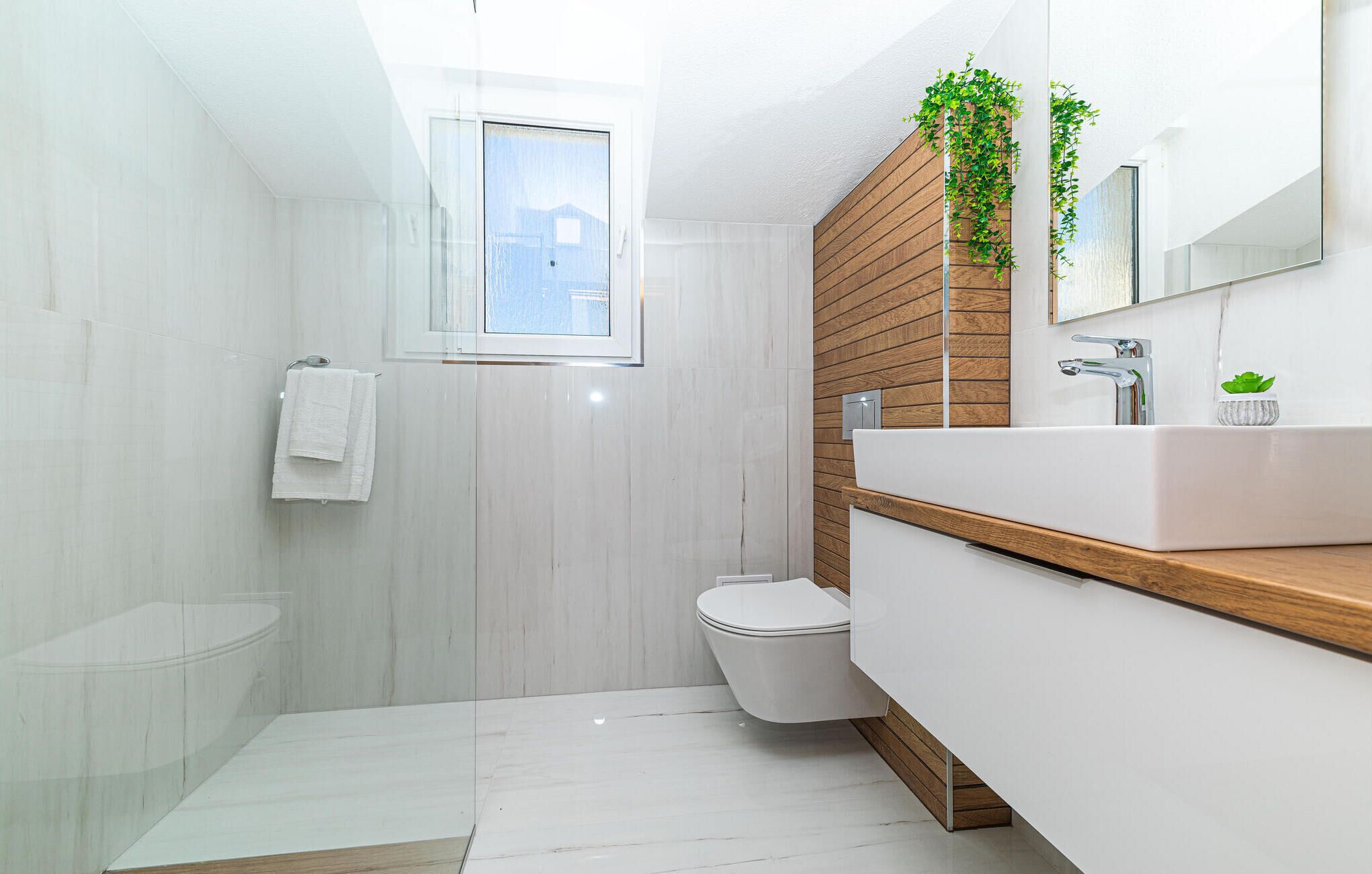 Un baño moderno con piso de mármol blanco, tocador de madera, inodoro blanco y ventana con vista al exterior.