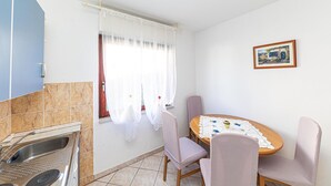 Une petite cuisine avec un évier, une fenêtre ornée de rideaux légers, une table à manger recouverte d’une nappe en dentelle et un tableau encadré accroché au mur.