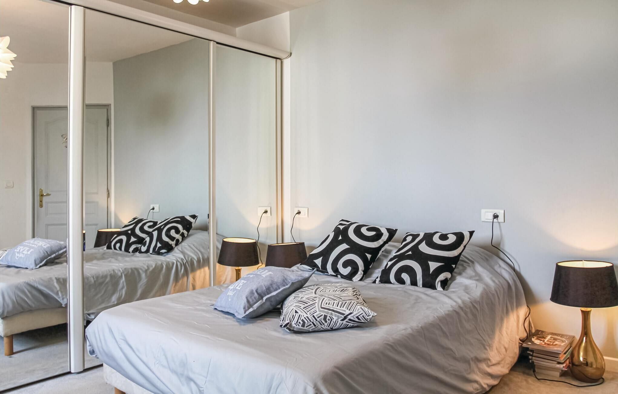 Un dormitorio moderno con un espejo grande, una cama con almohadillas estampadas, lámparas de noche y una mesita de noche con libros.
