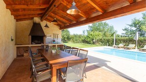 Cozinha externa com churrasqueira, mesa de jantar e cadeiras, com vista para uma piscina e jardim.