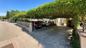 A covered parking area with a red car parked under a vine-covered pergola, adjacent to a building with a stone wall and a staircase leading to a door.
