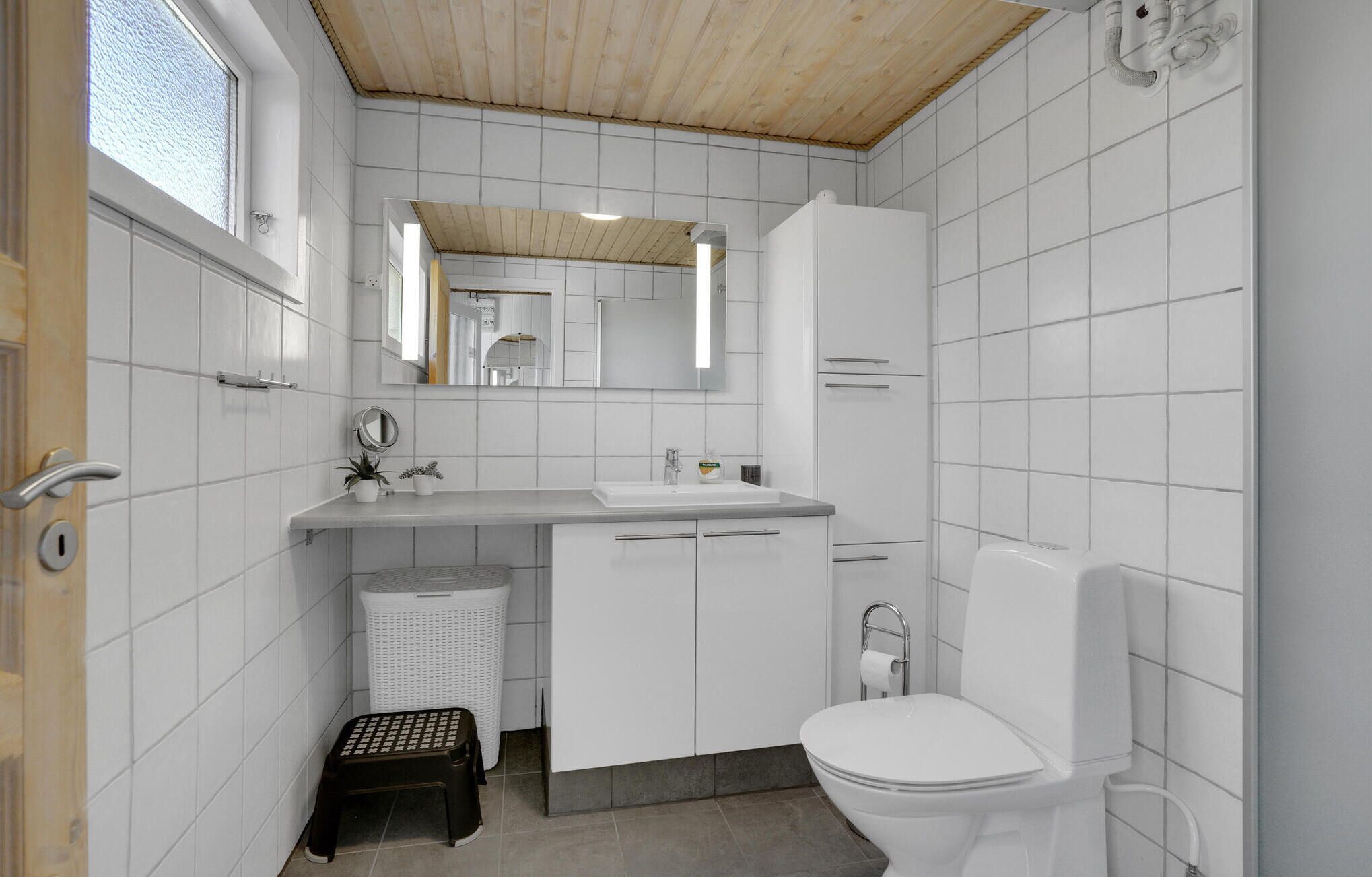 Ein modernes Badezimmer mit weißen Fliesen, Holzboden, einem Waschbecken mit Spiegel, einer Toilette und einer kleinen Stufe.
