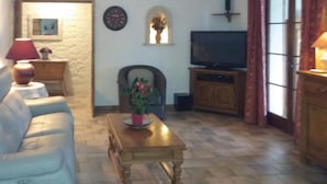 Un salón con sofá, una mesa de centro de madera con planta en maceta, un televisor sobre un mueble de madera, y una pared de piedra con un reloj y una ventana pequeña.