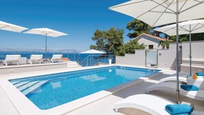 Una piscina con sillas de descanso y sombrillas, con vista a un cielo azul despejado y al mar.
