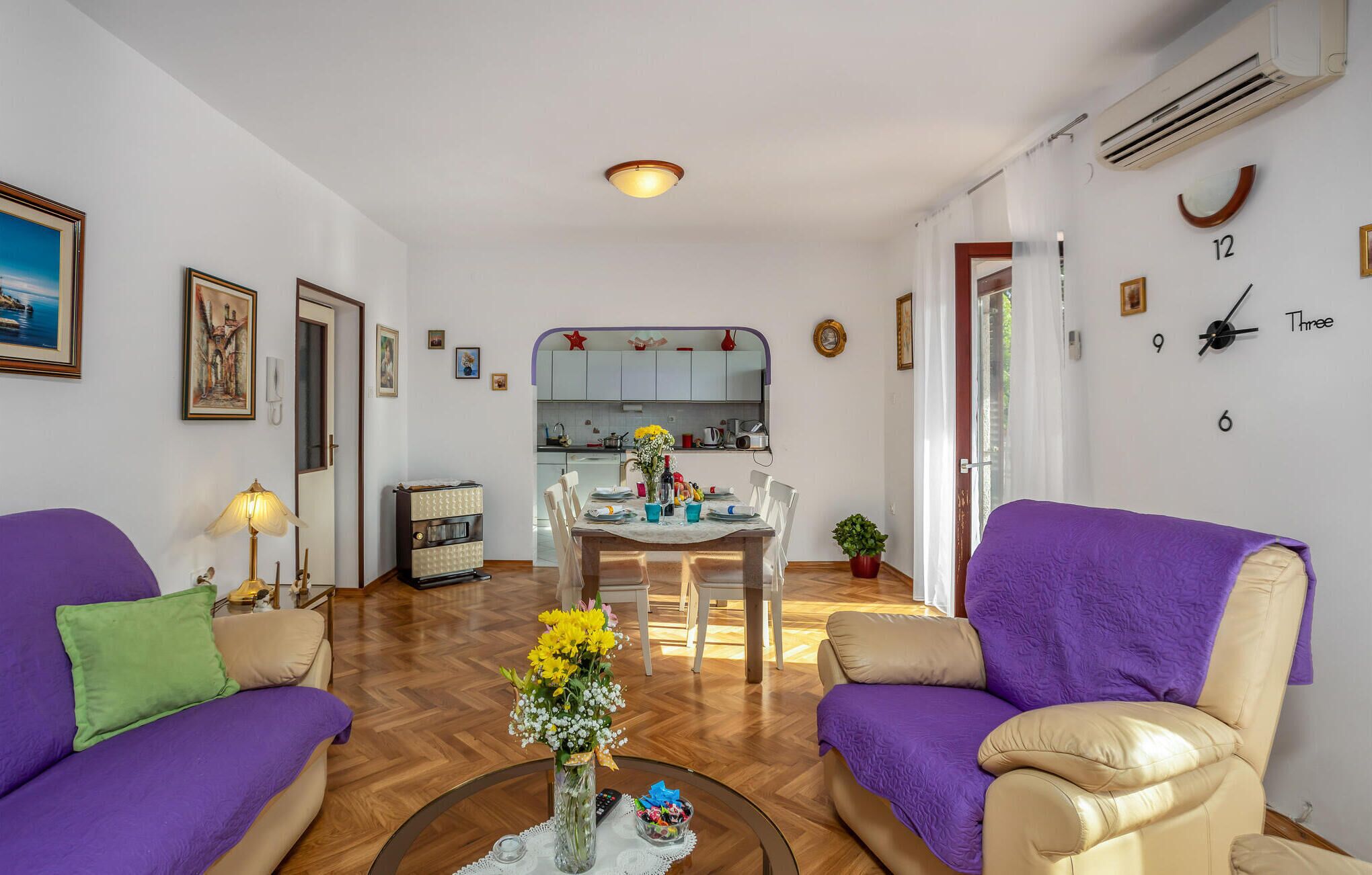 A living room with purple sofas, a coffee table with flowers, and a kitchen in the background.