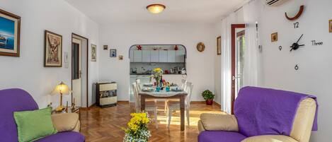 A living room with purple sofas, a coffee table with flowers, and a kitchen in the background.