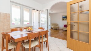 Un comedor con una mesa y sillas de madera, un frutero y vistas a través de una puerta abierta hacia una sala de estar con un sofá y un cuadro.