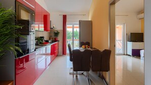 A modern kitchen with red cabinets, a dining area with a table and chairs, and a television in the adjacent room. - Lovely home in Betiga (Betiga)