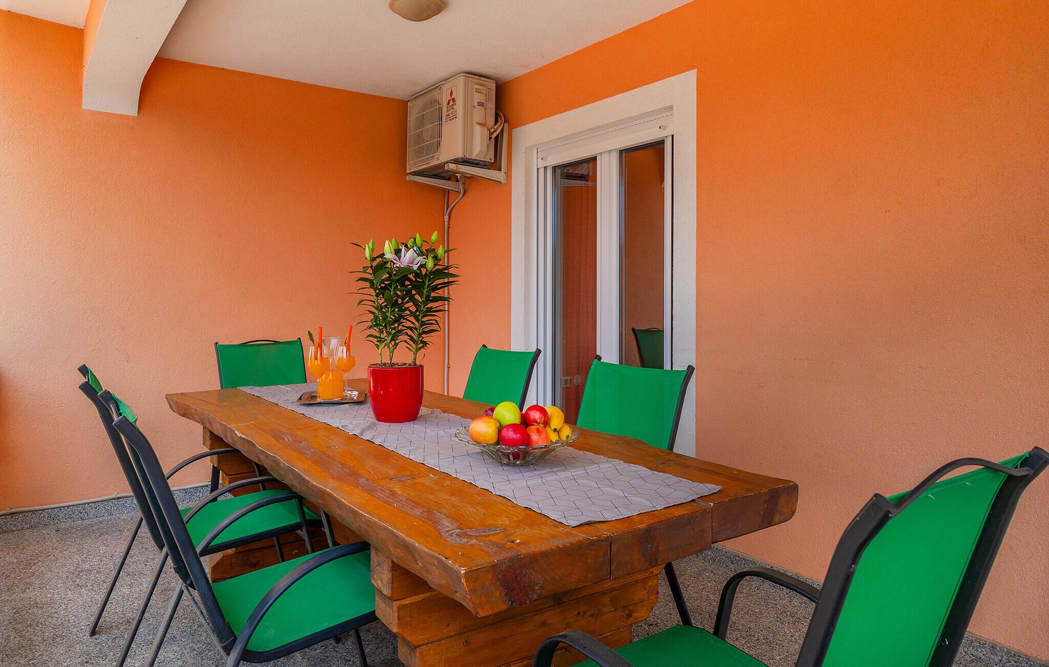 A wooden table with a glass top, surrounded by green chairs, a potted plant, and a fruit bowl.