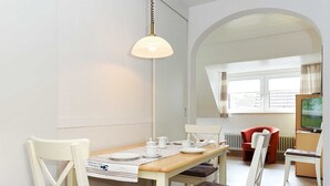 A dining area with a wooden table, white chairs, a hanging light fixture, and a television in the background.