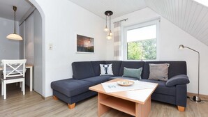 A modern living room with a dark blue sectional sofa, a wooden coffee table, and a white chair. There is a window with a view of greenery outside, and a pendant light hangs above the sofa.