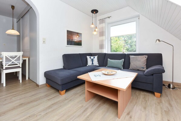 A modern living room with a dark blue sectional sofa, a wooden coffee table, and a white chair. There is a window with a view of greenery outside, and a pendant light hangs above the sofa.