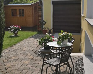 Une terrasse avec une table ronde et des chaises, entourée de plantes en pot et d’un abri de jardin en bois.