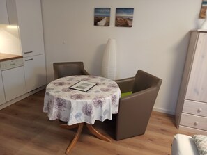 Un petit coin repas avec une table ronde recouverte d’une nappe à motifs floraux, deux chaises marron, un grand vase blanc et deux tableaux accrochés au mur.