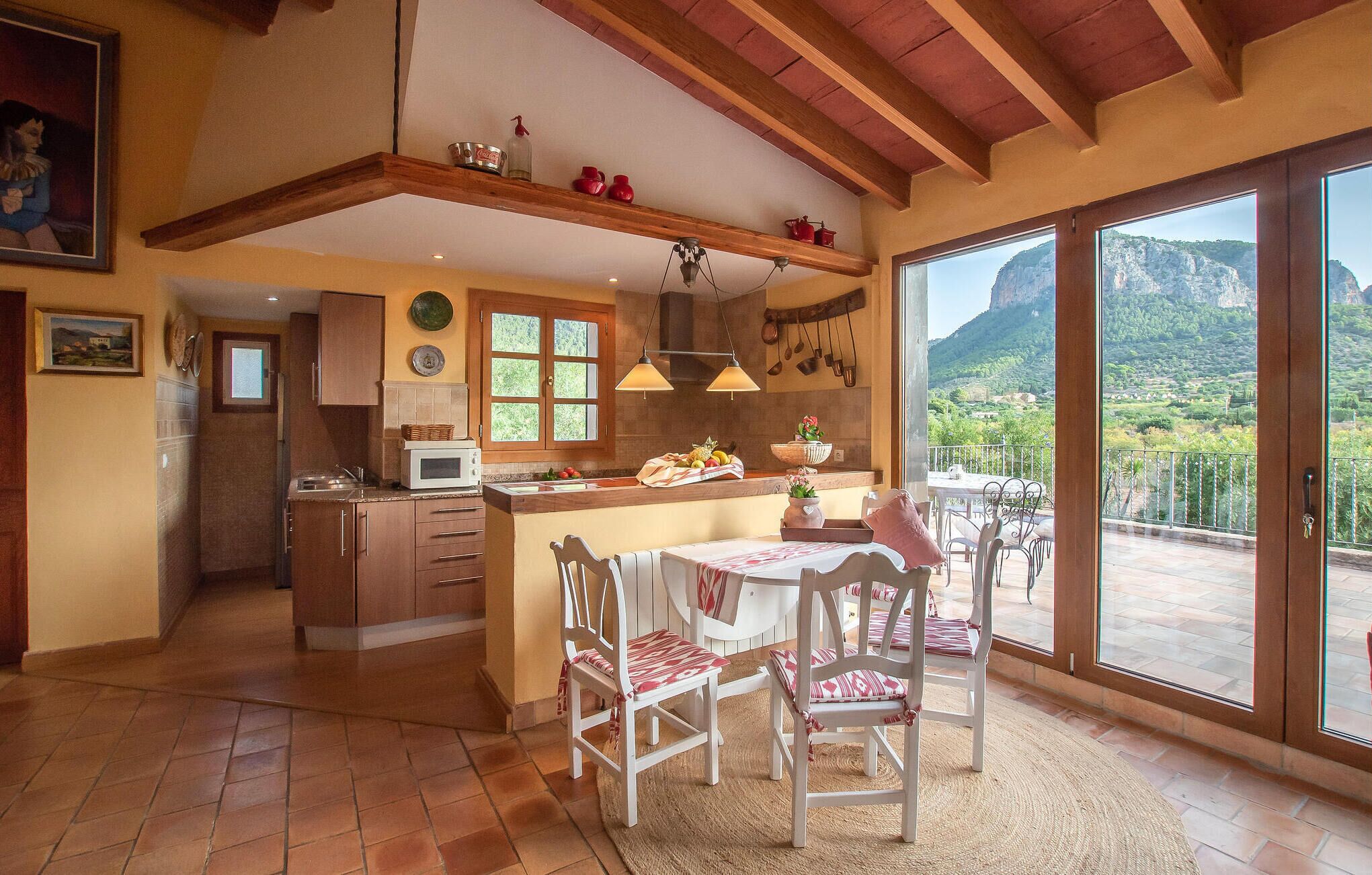 A spacious kitchen with wooden beams, a dining area with white chairs, and a large sliding glass door leading to a scenic mountain view.
