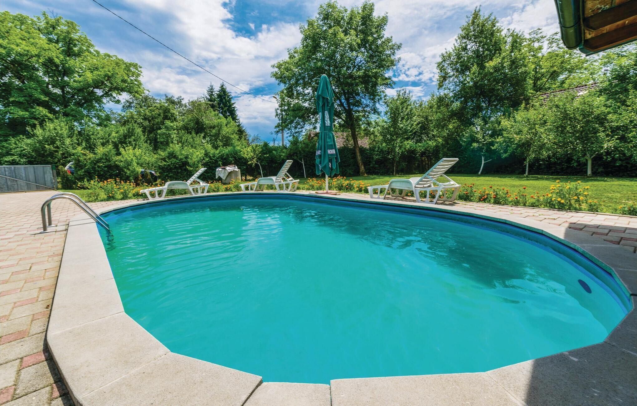 A rectangular swimming pool with a metal ladder on one side, surrounded by a brick deck. There are white lounge chairs and a green umbrella near the pool.